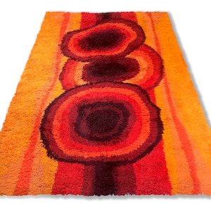 May include: A vintage shag rug with a geometric pattern of concentric circles in shades of orange, red, and brown. The rug has a long, rectangular shape and a thick pile.