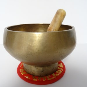 Pode incluir: Uma taça de canto de bronze com um malho de madeira dentro. A taça assenta numa almofada vermelha com acabamento dourado. A taça tem uma forma arredondada e uma superfície lisa e polida. A almofada tem cerca de 10 cm de diâmetro.