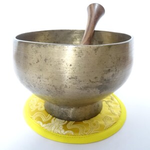 Pode incluir: Uma tigela de canto de cor bronze com um malho de madeira dentro, repousando sobre uma almofada amarela com um padrão floral. A tigela tem uma forma larga e arredondada e uma aparência ligeiramente envelhecida.