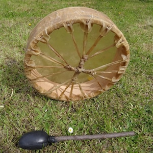 Könnte beinhalten: Eine handgefertigte Trommel mit einem Naturfell und Holzrahmen. Die Trommel ist beige und braun, mit einem dunkelbraunen Schlägel daneben. Die Trommel liegt auf grünem Gras.
