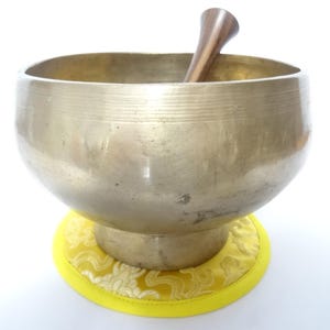 Pode incluir: Uma taça de canto de cor bronze com um malho de madeira no interior. A taça está sobre uma almofada amarela com um padrão floral. A taça tem uma forma arredondada e uma superfície lisa. O malho é castanho escuro.