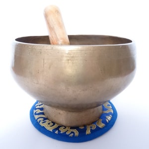 Pode incluir: Uma tigela de canto prateada com um batedor de madeira, descansando sobre uma almofada azul com bordados dourados.