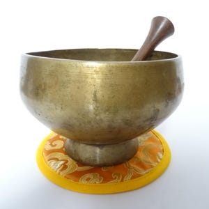 Pode incluir: Uma tigela de canto de cor bronze com um malho de madeira, repousando sobre uma almofada estampada amarela e laranja. A tigela tem formato arredondado e assenta sobre uma base de pedestal. O malho é castanho escuro.