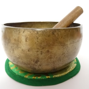 May include: A large, antique-looking, brass singing bowl with a wooden striker. The bowl is resting on a green and gold cushion.