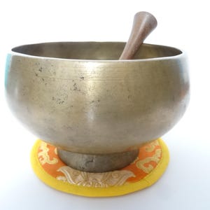 Pode incluir: Uma tigela de canto de cor bronze com um malho de madeira dentro, colocada sobre uma almofada com padrão laranja e amarelo. A tigela tem formato arredondado e superfície lisa.