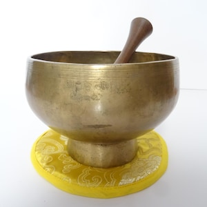 Pode incluir: Uma tigela de canto de bronze com um batedor de madeira, repousando sobre uma almofada amarela com um padrão decorativo. A tigela tem detalhes gravados e um acabamento polido. O batedor de madeira é marrom escuro.