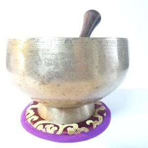 Pode incluir: Uma taça de canto de bronze com um batedor de madeira, repousando sobre uma almofada roxa e dourada. A taça tem marcações gravadas e uma forma arredondada. O batedor é castanho escuro. A almofada tem um padrão floral dourado.