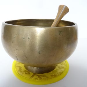 Pode incluir: Uma tigela de canto de latão com um batedor de madeira, descansando sobre uma almofada amarela com desenhos intrincados. A tigela tem uma forma arredondada e uma superfície lisa e polida.