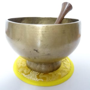 Pode incluir: Uma tigela de canto de cor bronze com um batedor de madeira repousa sobre uma almofada amarela com um padrão floral. A tigela é redonda com uma abertura larga, e o batedor é castanho escuro.