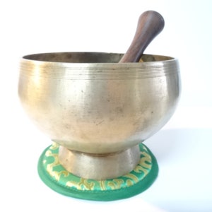 Pode incluir: Uma tigela de canto de latão com um batedor de madeira, descansando sobre um almofada verde e dourada. A tigela tem uma forma arredondada e uma borda larga.