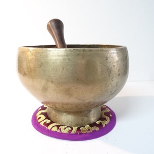 Pode incluir: Uma tigela de canto de latão com um batedor de madeira, descansando sobre um almofada roxa com bordados dourados. A tigela tem uma forma arredondada e uma borda larga.