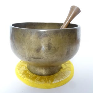 Pode incluir: Uma tigela de canto de bronze com um batedor de madeira repousa sobre uma almofada amarela com um design ornamentado. A tigela tem uma forma arredondada e uma aparência lisa e envelhecida, sugerindo seu uso em meditação ou cura sonora.