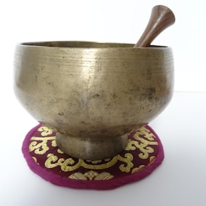 Pode incluir: Uma tigela de canto de cor bronze com um batedor de madeira repousa sobre uma almofada redonda roxa com filigrana dourada. A tigela tem uma forma arredondada e uma superfície texturizada. O batedor de madeira é curvo e afilado.