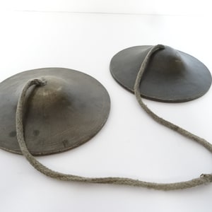 May include: Two small, round, gray metal cymbals connected by a long, gray rope. The cymbals are slightly concave and have a smooth surface.