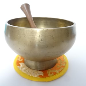 Pode incluir: Uma taça de canto de cor bronze com um malho de madeira no interior, repousando sobre uma almofada com padrão laranja e amarelo. A taça tem formato arredondado e base. O malho é castanho claro.