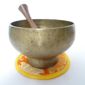 Pode incluir: Uma tigela de canto de cor bronze com um malho de madeira dentro, colocada sobre uma almofada estampada amarela e laranja. A tigela e o malho estão sobre um fundo branco.