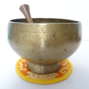 Pode incluir: Uma tigela de canto de cor bronze com um malho de madeira dentro. A tigela está sobre uma almofada estampada amarela e laranja. A tigela tem marcações gravadas.
