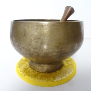 Pode incluir: Uma tigela de canto de cor bronze com um malho de madeira, repousando sobre uma almofada amarela com um padrão dourado. A tigela tem formato arredondado e uma abertura larga. O malho é marrom.