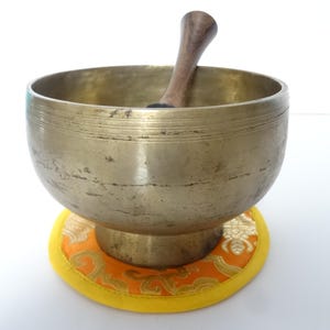 Pode incluir: Uma tigela de canto de bronze com um batedor de madeira repousa sobre uma almofada estampada amarela e laranja. A tigela tem formato arredondado com uma abertura larga e um batedor de madeira escura. A almofada tem um desenho floral dourado.