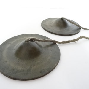 May include: Two small, antique bronze-colored finger cymbals connected by a twisted rope. The cymbals have a slightly concave shape with a central raised area. The rope is a light gray color.