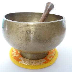 Pode incluir: Uma tigela de canto de bronze com um batedor de madeira dentro. A tigela repousa sobre uma almofada com estampa amarela e laranja. A tigela tem formato arredondado e aparência lisa e envelhecida, sugerindo seu uso em meditação ou cura sonora.