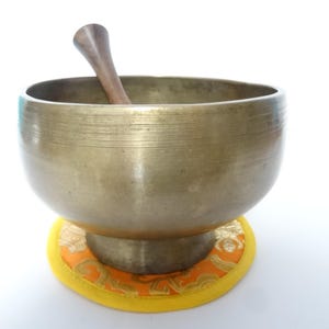 Pode incluir: Uma tigela de canto de bronze com um batedor de madeira repousa sobre uma almofada estampada em laranja e amarelo. A tigela tem uma forma larga e arredondada e um acabamento metálico liso. O batedor de madeira é curvo e marrom escuro.