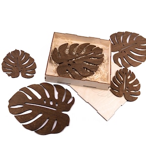 May include: A set of wooden coasters in the shape of Monstera leaves. The coasters are a dark brown color and are displayed with a wooden box and a wooden lid. The coasters are designed to protect surfaces from heat and moisture.