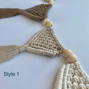 May include: A macrame garland with beige and white triangle flags and wooden beads. The garland has a tassel on each flag. The text "Style 1" is visible in the image.