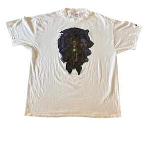 May include: White t-shirt featuring a graphic print of a man holding a glowing staff, with two faces in the background. The graphic is in shades of blue, green, and black. The shirt is a standard crew neck.