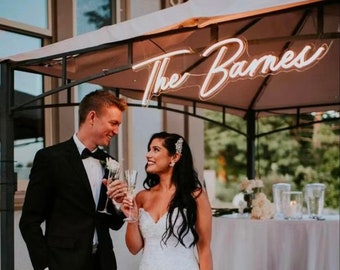 May include: A bride and groom at a wedding ceremony, smiling and raising a toast with champagne flutes. A neon sign above them reads "The Barnes".