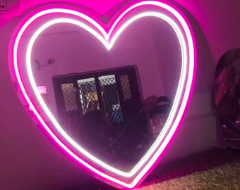 Op de afbeelding: Een grote hartvormige spiegel met een dubbele neon roze en witte omtrek. De spiegel hangt aan een roze muur en reflecteert een kamer met een raam en meubels.
