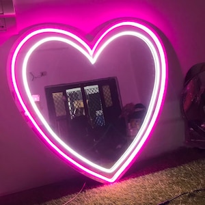 Puede incluir: Un gran espejo con forma de corazón con un doble contorno de neón rosa y blanco. El espejo está colgado en una pared rosa y refleja una habitación con una ventana y muebles.
