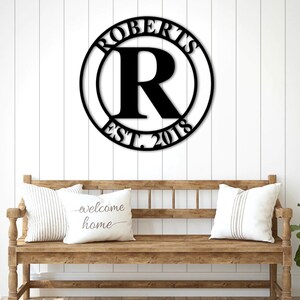 Op de afbeelding: Zwart metalen bord met de achternaam "Roberts" en het jaar "2018" in een cirkel met een grote "R" in het midden. Het bord hangt aan een witte muur boven een houten bank met kussens.