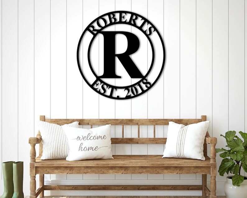 Op de afbeelding: Zwart metalen bord met de achternaam "Roberts" en het jaar "2018" in een cirkel met een grote "R" in het midden. Het bord hangt aan een witte muur boven een houten bank met kussens.
