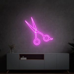 Puede incluir: Un letrero de neón rosa brillante con forma de tijeras de peluquería. El letrero está montado en una pared gris oscuro sobre un mueble gris. Las tijeras son un elemento decorativo para una peluquería.