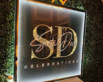 May include: A black and gold sign with the text "SD Secta Devi CELEBRATIONS" in a gold glitter font. The sign is illuminated with white lights and is framed with a silver frame.