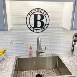 Op de afbeelding: Een zwart metalen bord met de tekst "Bennett Est. 2021" en de letter "B" in een cirkel. Het bord is bevestigd aan een witte betegelde muur boven een roestvrijstalen gootsteen.