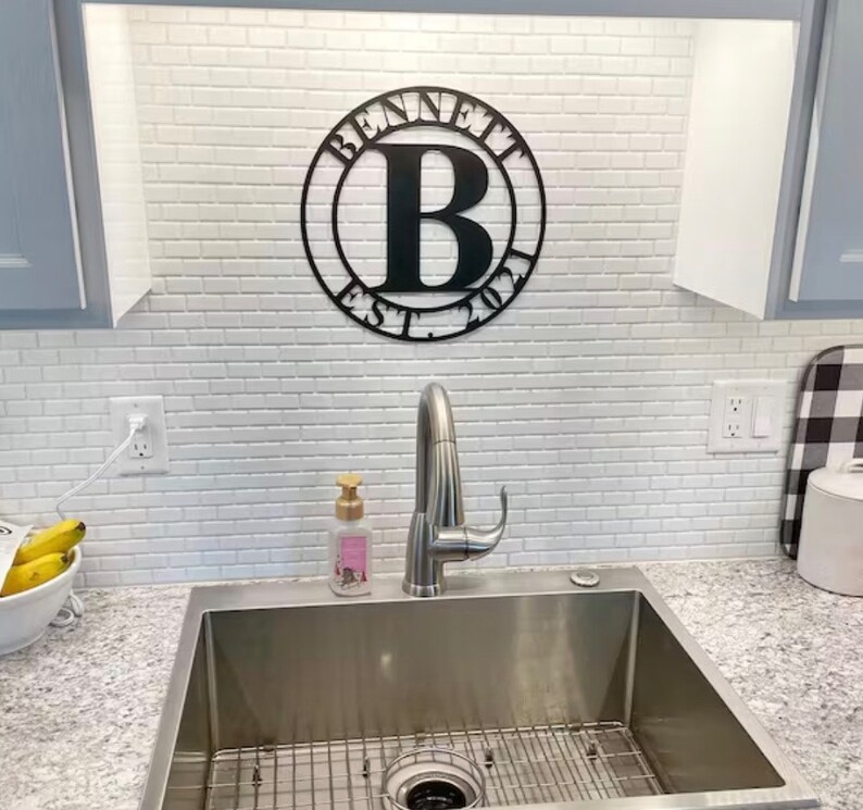 Op de afbeelding: Een zwart metalen bord met de tekst "Bennett Est. 2021" en de letter "B" in een cirkel. Het bord is bevestigd aan een witte betegelde muur boven een roestvrijstalen gootsteen.