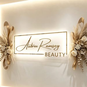May include: A rectangular wall sign with a white backlit panel featuring the gold text Aubree Ramsey BEAUTY. The sign is flanked by two decorative arrangements of dried pampas grass, eucalyptus leaves, white roses, and curved gold metal accents.