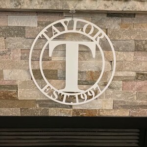 Op de afbeelding: Witte metalen cirkel met het woord "TAYLOR" en "EST. 1991" eromheen en een grote letter "T" in het midden.