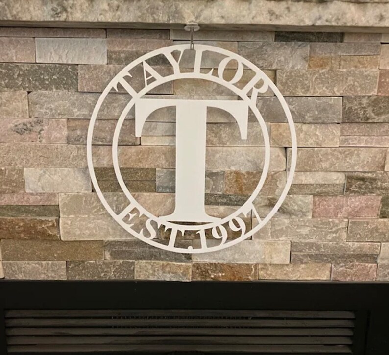Op de afbeelding: Witte metalen cirkel met het woord "TAYLOR" en "EST. 1991" eromheen en een grote letter "T" in het midden.