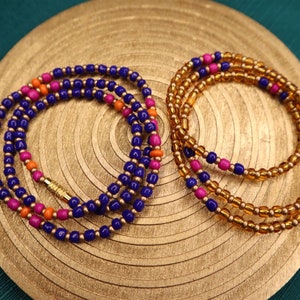 May include: Two beaded necklaces. One is navy blue with pink and orange beads, the other is gold with pink and blue beads. The necklaces are on a gold-colored, textured surface with a green background.