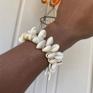 Puede incluir: Un brazalete hecho de conchas marinas blancas y crema. Las conchas están ensartadas para formar un brazalete tipo puño. El brazalete se usa en un brazo de piel oscura.