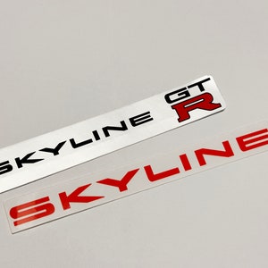 Peut inclure: Deux autocollants blancs avec du texte noir qui dit "SKYLINE" et "GT-R". L'autocollant "GT-R" a un "R" rouge. Il y a aussi un autocollant transparent avec du texte rouge qui dit "SKYLINE".