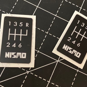 Op de afbeelding: Twee zwart-witte NISMO-schakelpatroon stickers. De stickers hebben een zwarte achtergrond met witte nummers en een wit NISMO-logo.