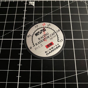 May include: A silver NISMO racing radiator cap with a black and red gauge and the text "RACING RADIATOR CAP" and "DANGER".