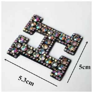 May include: A black iron-on patch in the shape of the letter H, covered in iridescent rhinestones. The patch measures 5.3 cm wide and 5 cm tall.