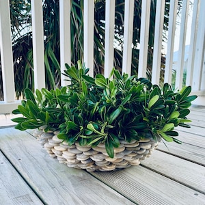 May include: A decorative planter filled with vibrant green foliage. The planter is constructed from small, smooth, gray stones, arranged in a layered pattern. The arrangement sits on a weathered wooden deck, with a white railing in the background.