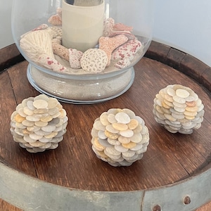 May include: Three small decorative spheres made of stacked smooth stones in shades of white, gray, and beige. The spheres are arranged on a wooden surface with a metal band.