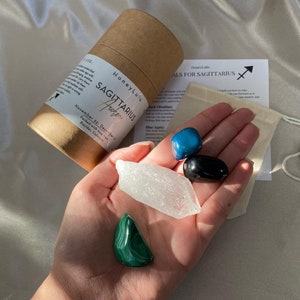 May include: A set of four crystals for the Sagittarius zodiac sign. The crystals are blue agate, black obsidian, clear quartz, and malachite. The crystals are arranged in a hand, with a small brown paper bag and a card with the text "Crystals for Sagittarius" in the background.